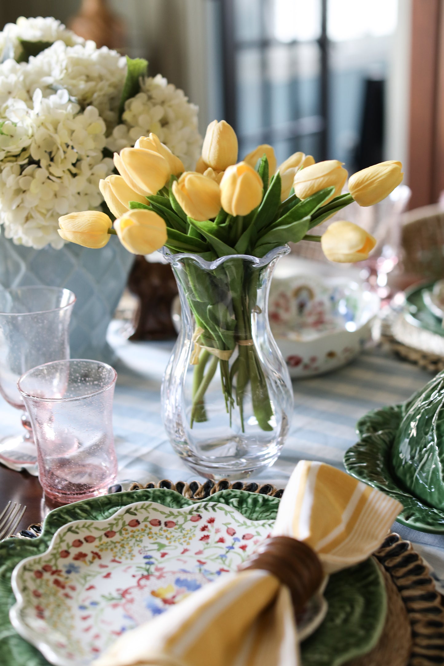 Decorative table setting with yellow tulips in a vase, floral plates, and striped tablecloth.