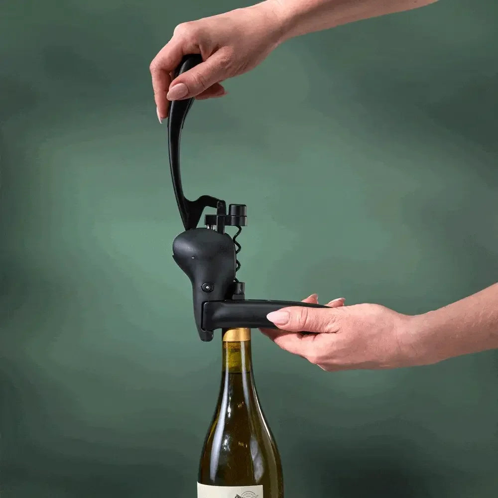 Hands using a black lever-style corkscrew to open a wine bottle against a green background
