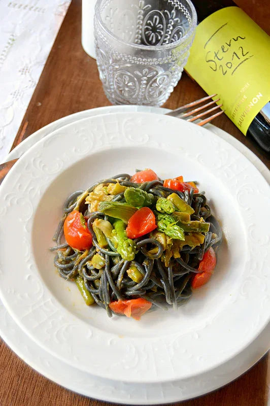 Black squid ink pasta with cherry tomatoes and green vegetables on white plate, served with a glass and white wine bottle