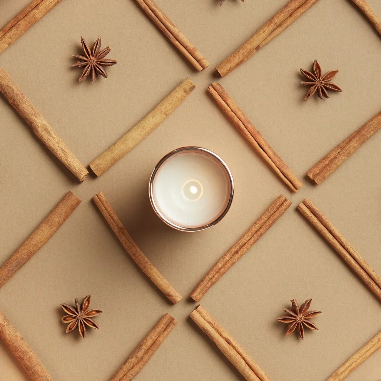 White votive candle surrounded by cinnamon sticks and star anise in a geometric pattern on beige background