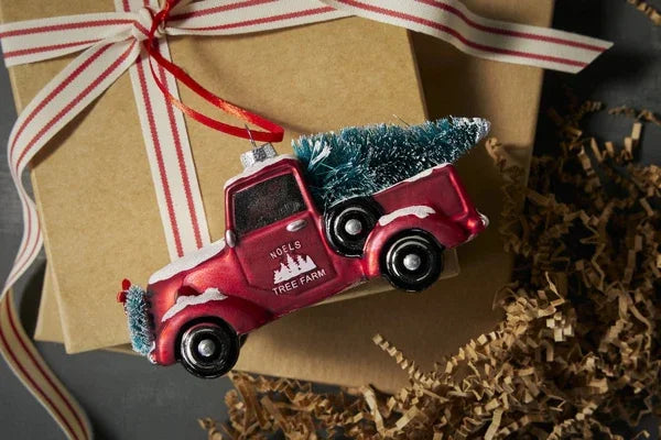 Red vintage truck ornament with a Christmas tree on back, labeled Noels Tree Farm, over gift boxes and shredded paper