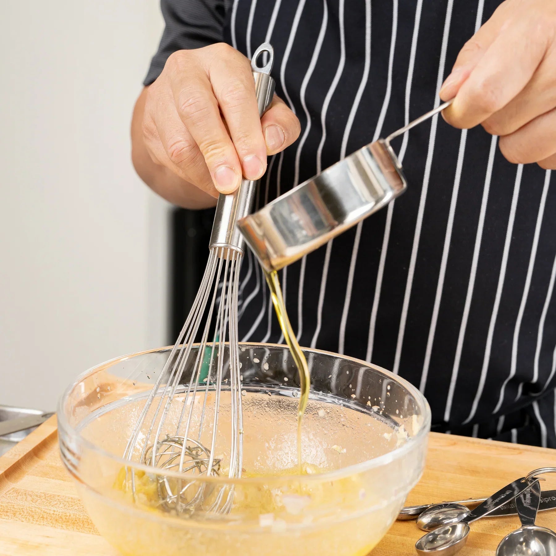 Person in striped apron whisking batter in glass bowl while pouring oil in kitchen