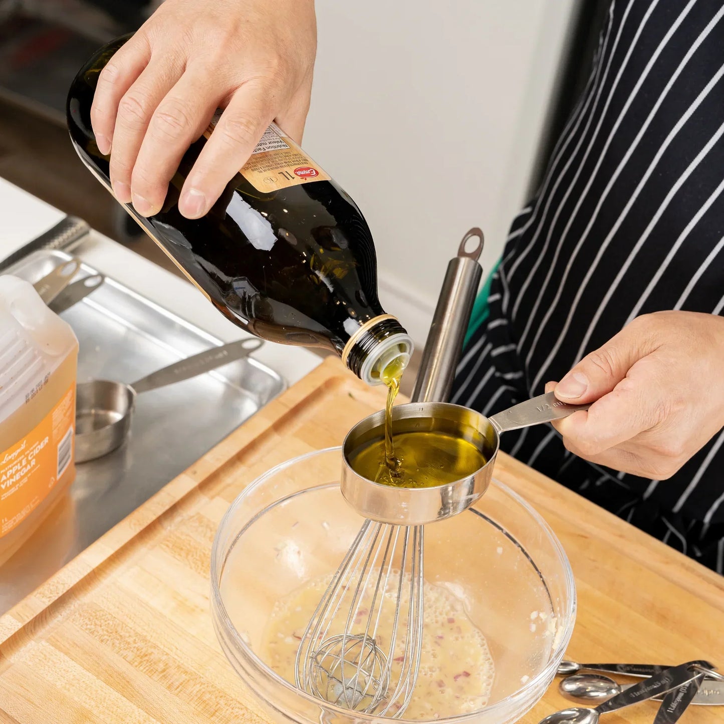 Person pouring olive oil into a stainless steel measuring cup over glass bowl with whisk on wooden cutting board