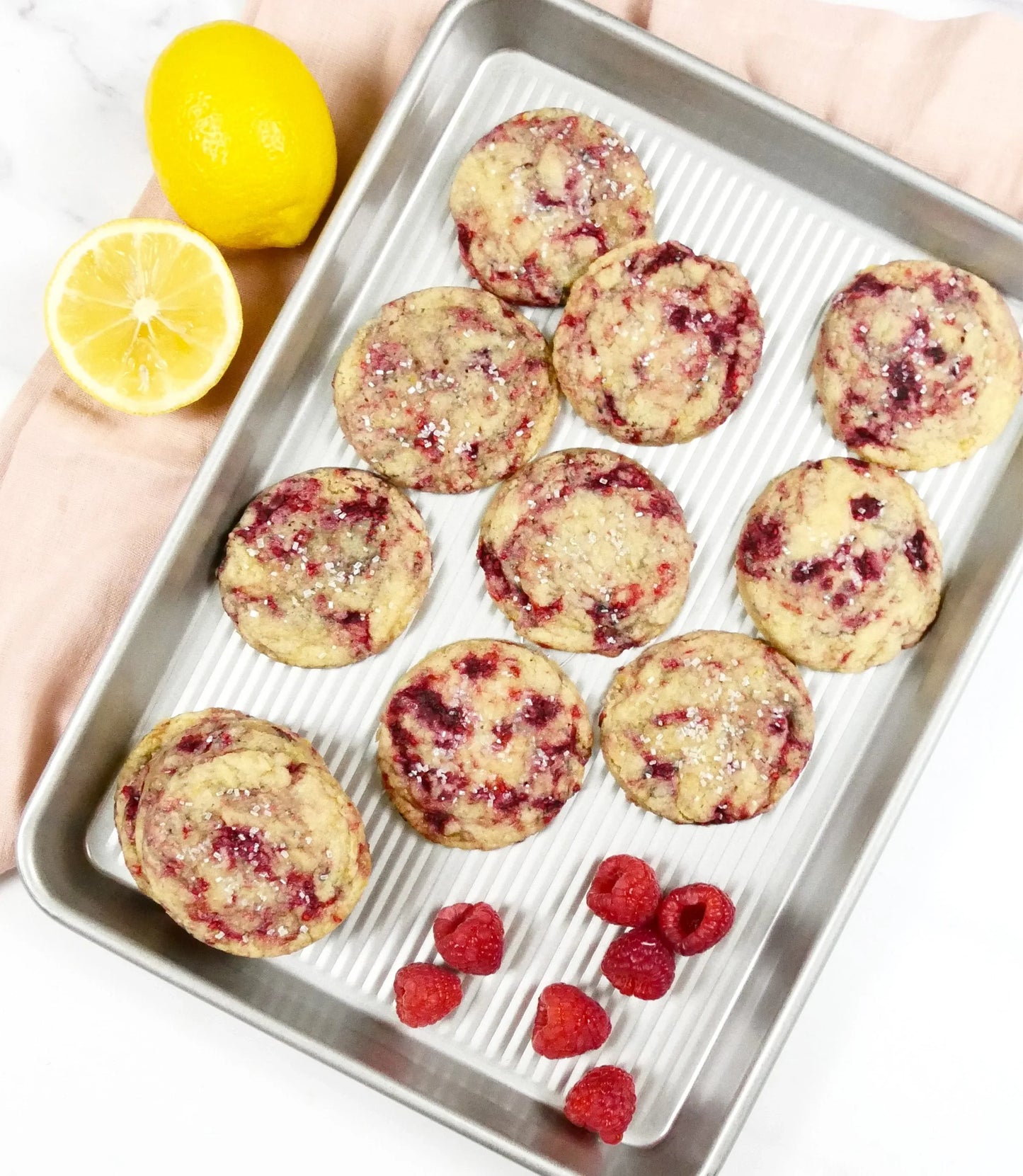 Raspberry lemon cookies with sugar sprinkles on a silver baking tray, fresh raspberries and lemons beside