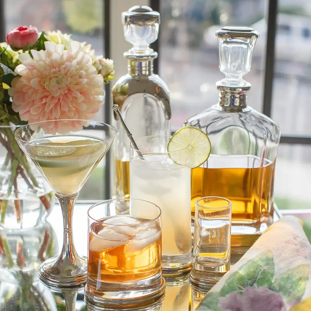 Assorted cocktails with ice, lime garnish, and whiskey decanters near a window and floral centerpiece
