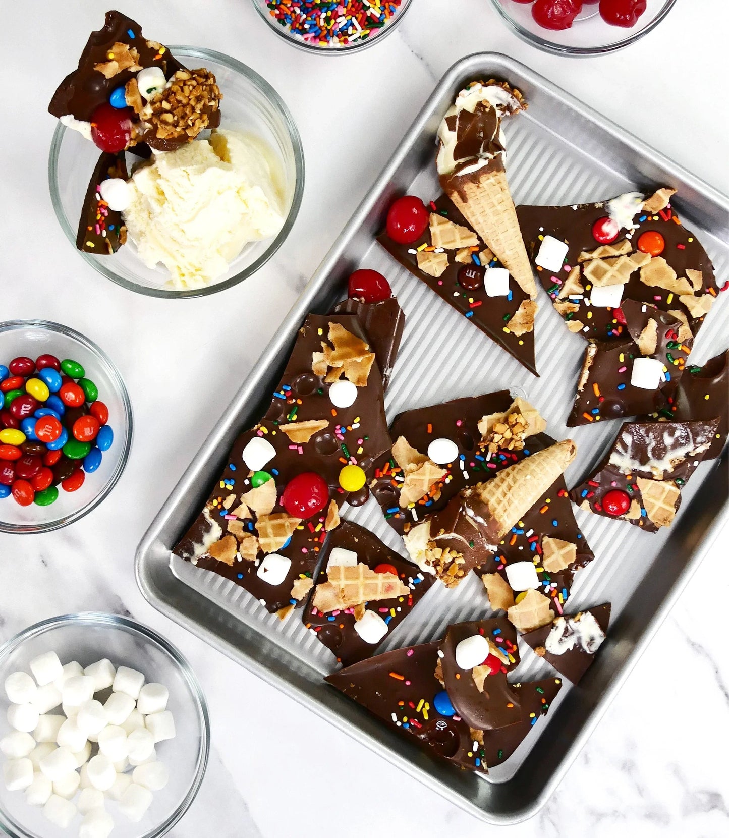 Chocolate bark with sprinkles, cherries, marshmallows, and waffle cone pieces on a baking sheet with ice cream and candy bowls