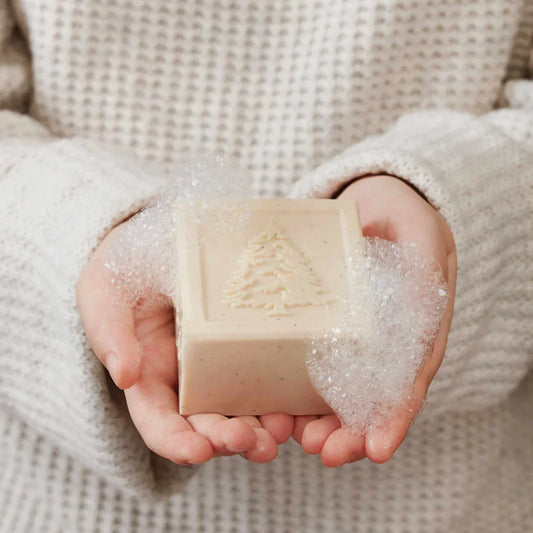Hands holding a beige bar soap with embossed pine tree and bubbles, wearing a knitted sweater