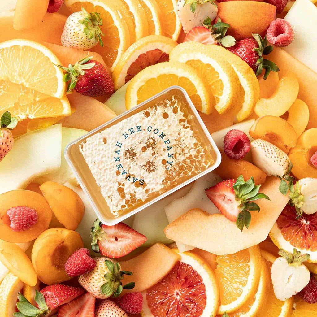 Honeycomb in a plastic container labeled Savannah Bee Company surrounded by fresh sliced oranges, strawberries, raspberries, and apricots