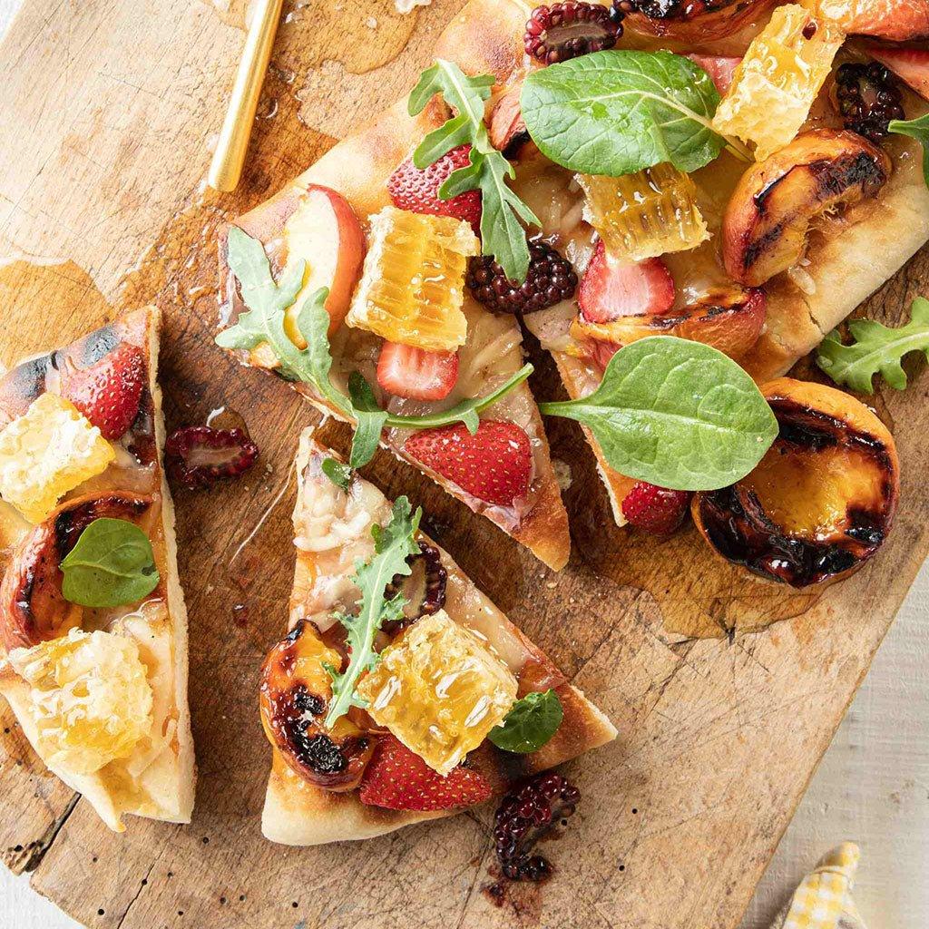 Slices of gourmet flatbread topped with honeycomb, strawberries, grilled peaches, spinach, arugula, and blackberries on a wooden board