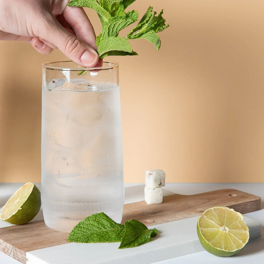 Hand garnishing mojito cocktail in glass with fresh mint leaves, lime halves, sugar cubes on wooden board