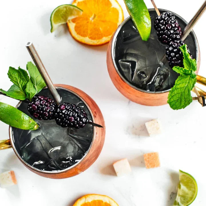 Two copper Moscow mule mugs with blackberry, lime, and mint garnish on a white surface with orange slices and sugar cubes