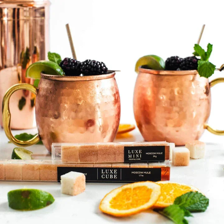 Copper mugs with Moscow Mule cocktails garnished with blackberries, lime, and mint beside Luxe Cube sugar cubes and orange slices