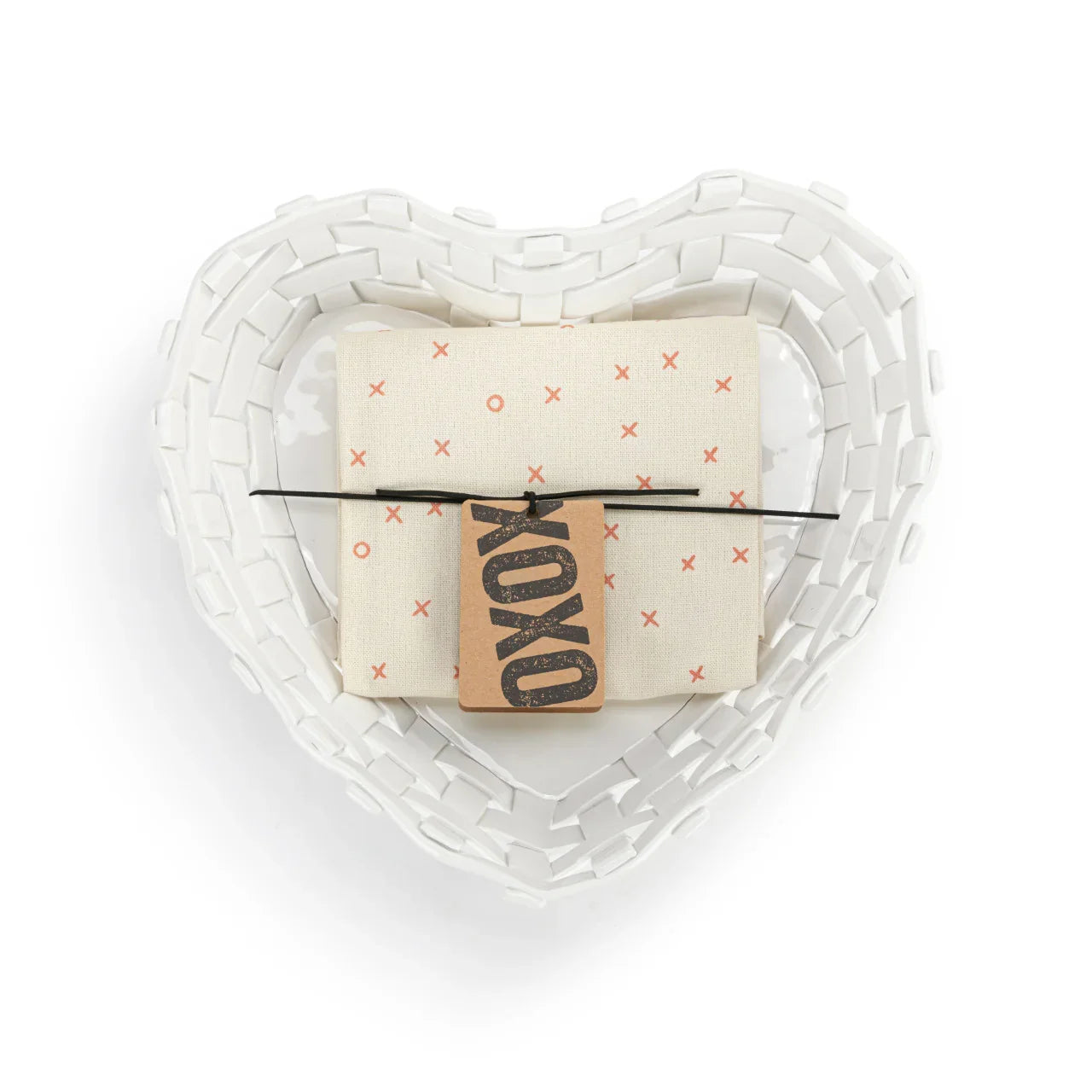 White woven heart-shaped basket holding a folded cloth with red X and O pattern and a brown tag reading XOXO
