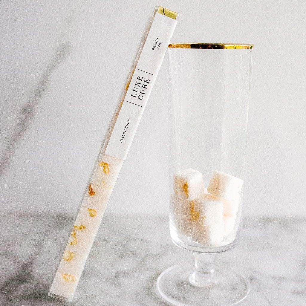 Clear glass with gold rim containing peach Luxe Cube sugar cubes next to tube packaging on marble surface
