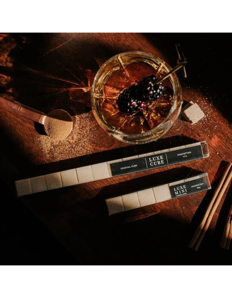 Manhattan cocktail with blackberries on a skewer, brown sugar cubes, cinnamon sticks, and Luxe Cube packaging on wooden surface