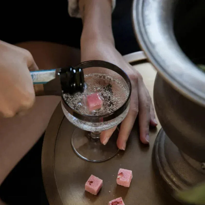 Hand pouring sparkling drink into coupe glass with pink tea cube on wooden table