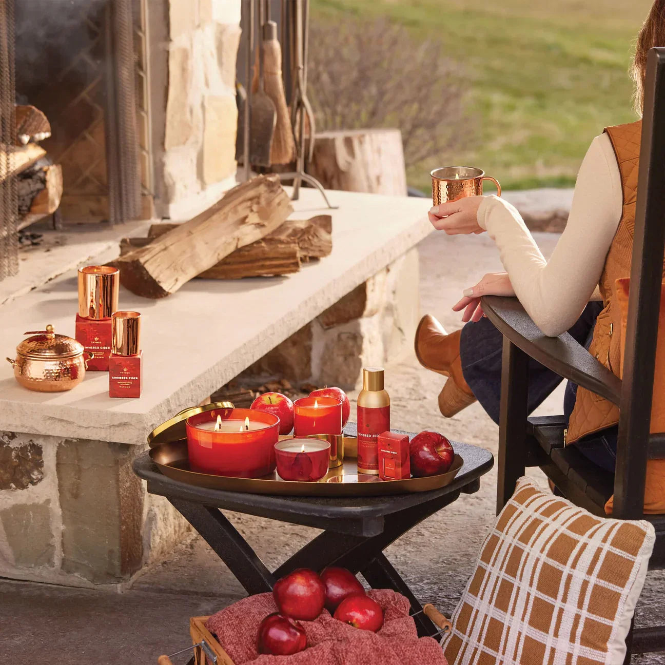 Outdoor cozy scene with simered cider candles, red apples, and a woman holding copper mug by fireplace